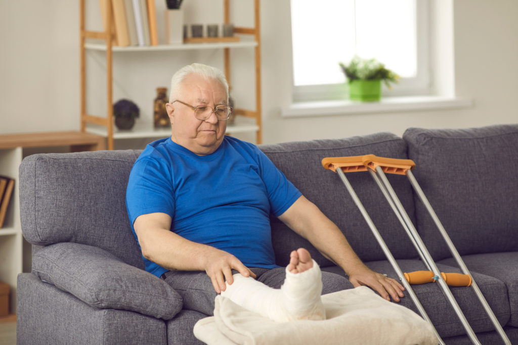 Older man with a leg cast sitting on a couch beside a pair of crutches
